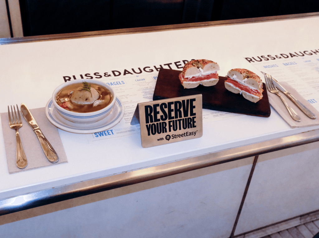 Image » new classic Soup in a bowl with a matzo ball and dill, accompanied by two bagel-sandwich halves on a wooden board; ’Reserve Your Future’ sign nearby on a deli counter table.