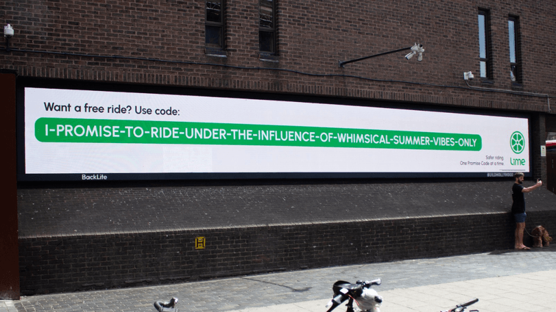 Digital billboard on a brick wall advertising a ride code in a green banner; a person and a dog stand to the right.