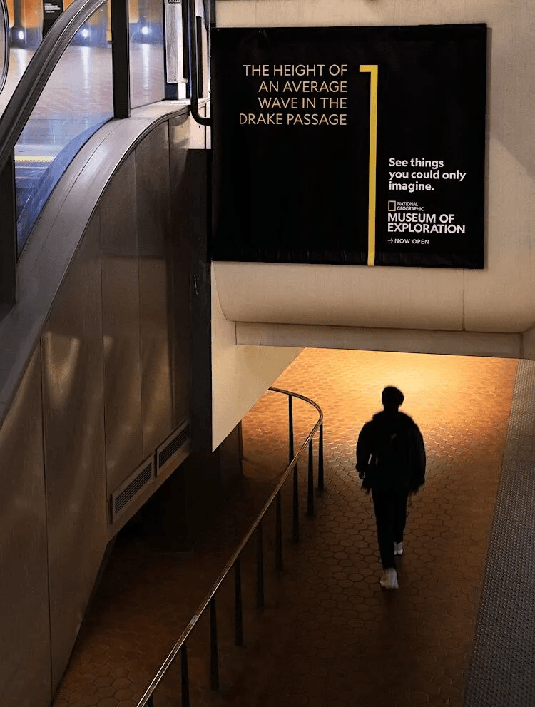 Image – new classic Person wearing a backpack walks down a sloped path in a modern transit area, past a large black poster advertising the National Geographic Museum of Exploration.